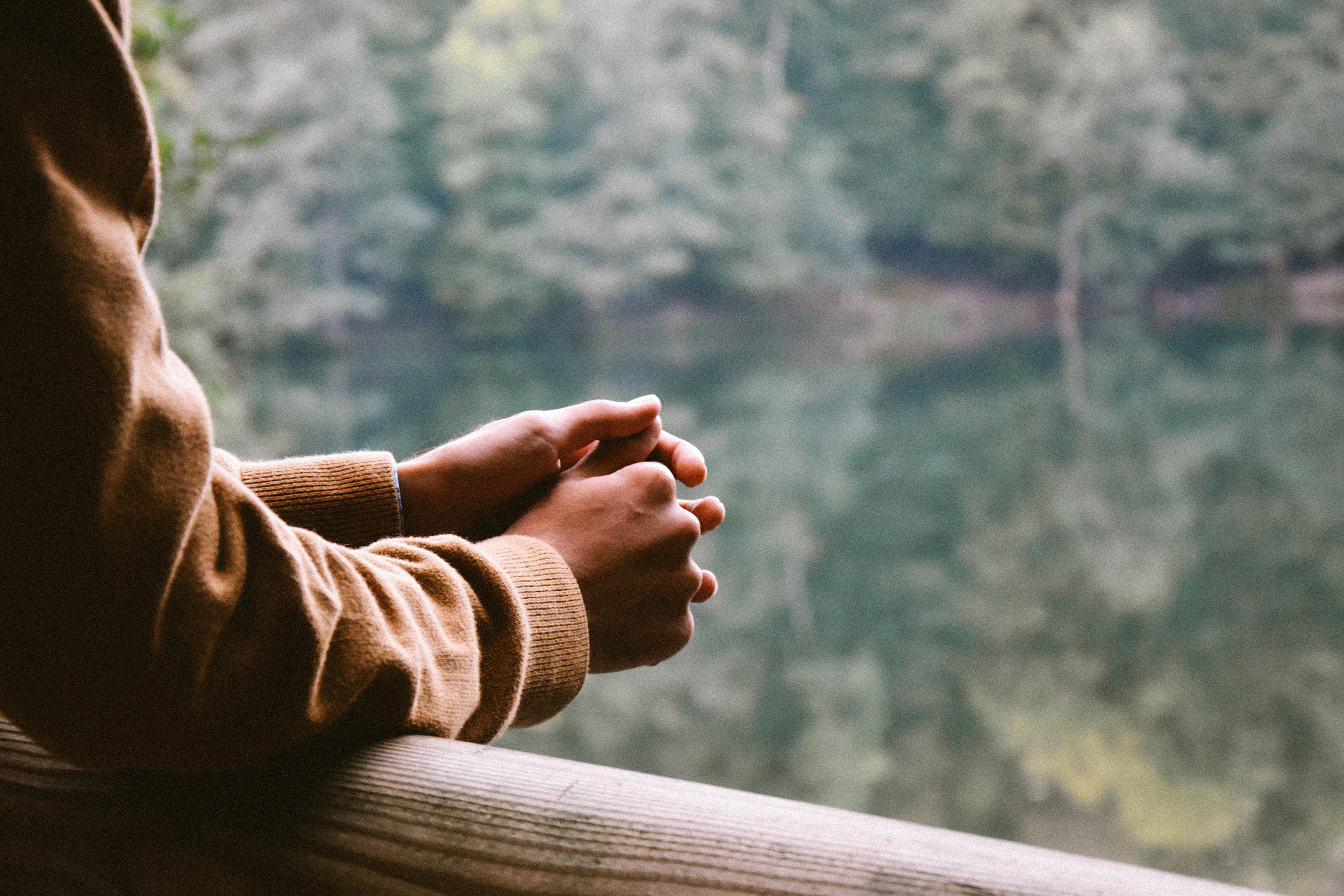 Close-up photo of the hands of a person leaving over a deck overlooking a lake surrounded by trees.