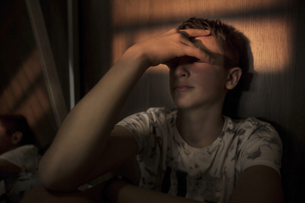 Teenager sitting in a dimly lit room with a hand across his forehead.