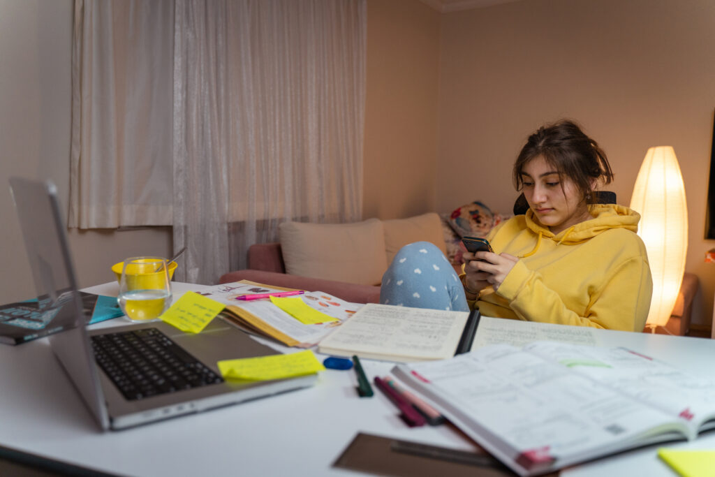 Teenager holding a smartphone at a desk with open notebooks and a laptop.