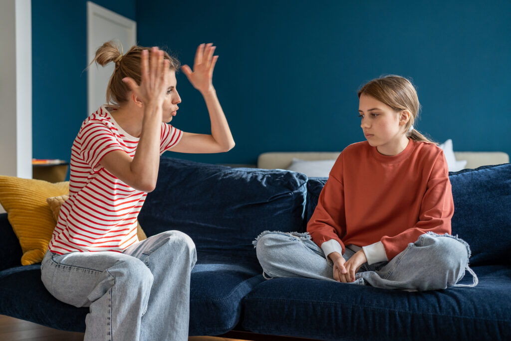 Mother raising her hands while speaking to her teenage daughter on a sofa at home.