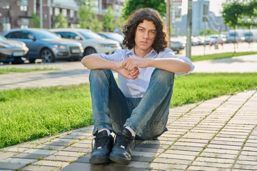 Teenage boy sitting on a paved path beside grass in a city park.