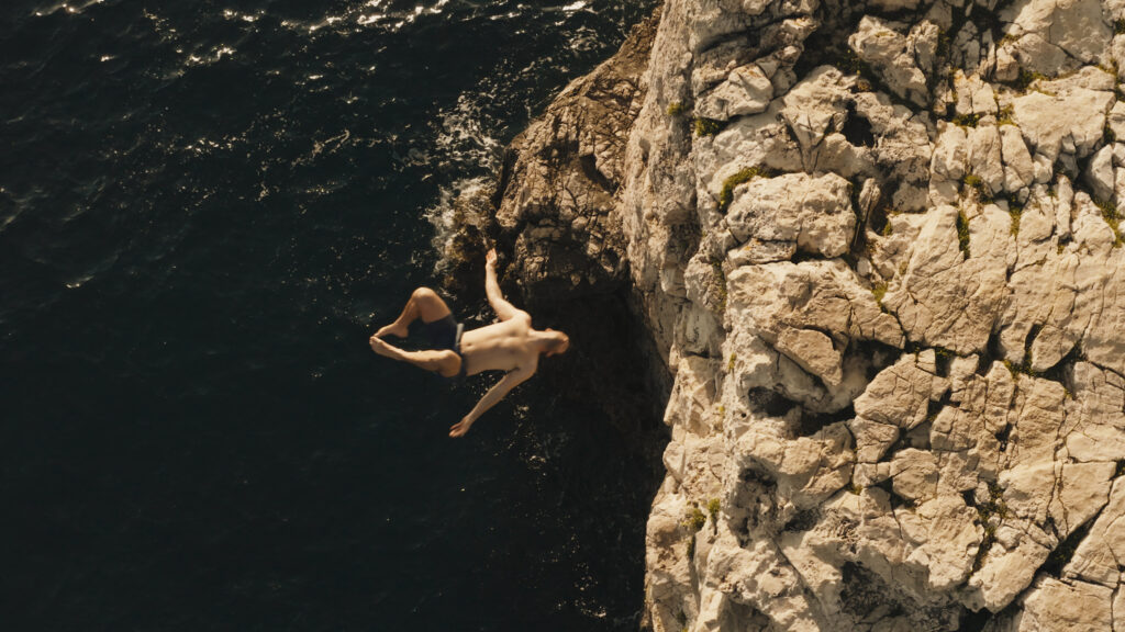 Young man mid-backflip above the sea beside a rocky cliff.