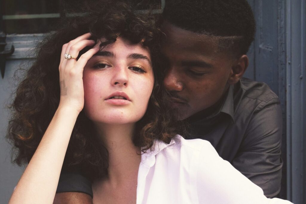 A woman with curly dark hair in a white shirt looks at the camera while a man in a dark shirt holds her from behind, his face turned toward her.