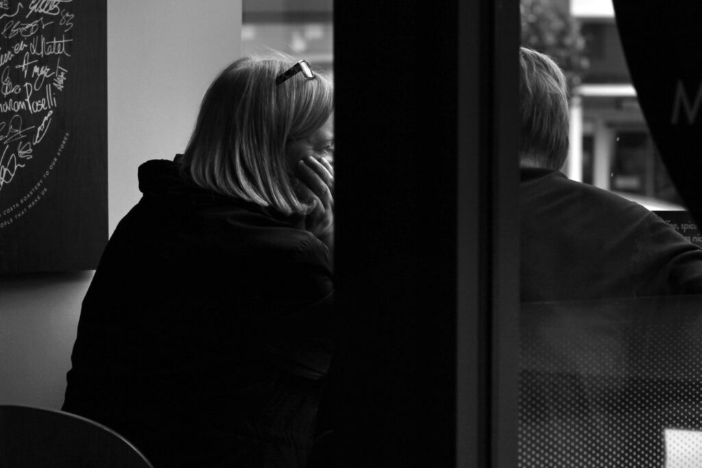 Emotional cheating: a woman alone at a coffee-shop table, hand against her cheek, behind a dark pillar