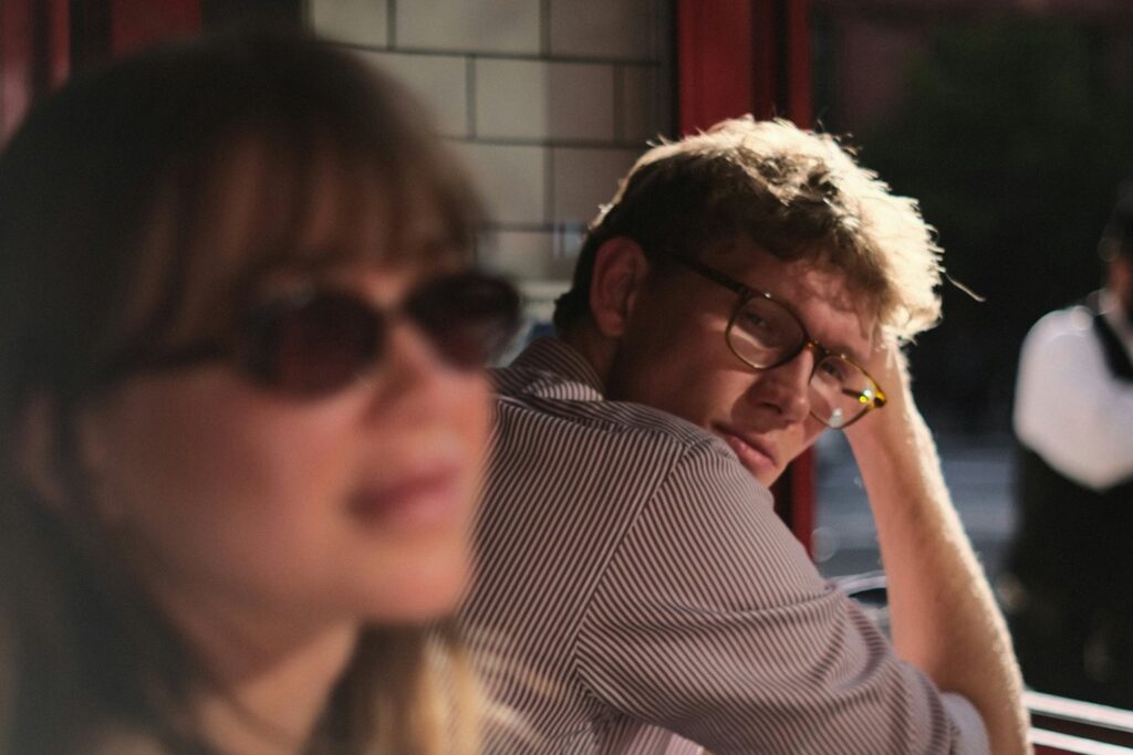 A couple at a cafe window: she looks past the camera in sunglasses while he leans on the counter and looks toward her, the kind of moment a partner can read as emotionally unavailable.