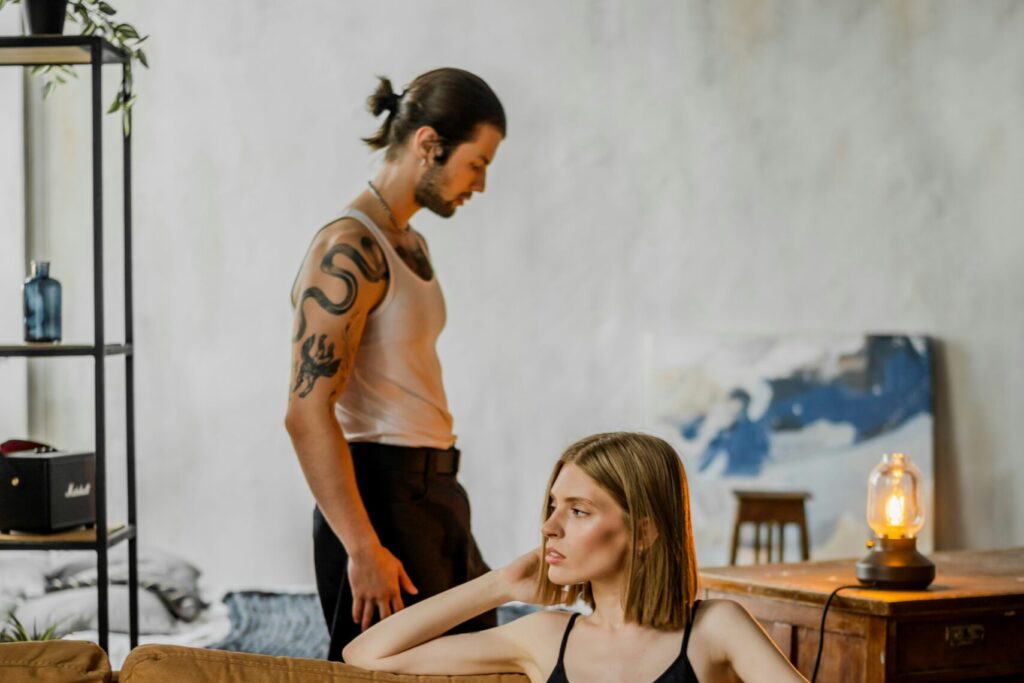 A woman sits on a mustard-colored sofa while a man stands behind her.
