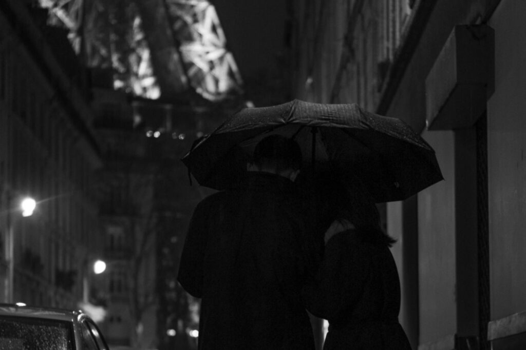 A couple walking close together under one umbrella on a rainy Paris street at night.