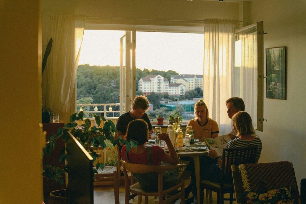Four people at a dinner table by an open window at sunset, one couple looking at a book together while the other two sit apart
