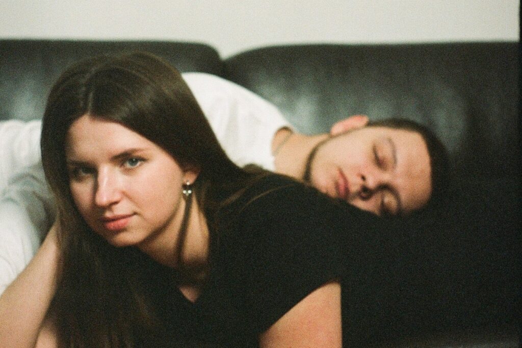 Situationship: a woman looks into the camera while a man sleeps against her back on a dark sofa
